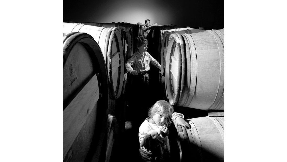 Portrait de Jean-Marie Gros, Château Grange Neuve (Pomerol - Cru classique d'Yvon Mau), et deux enfants entre deux rangées de fûts dans un chai ; © Richard Nourry